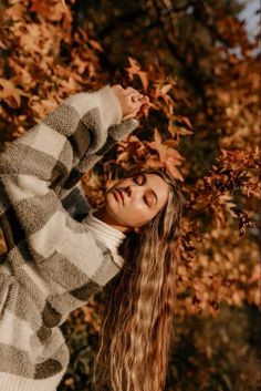 a woman with long hair standing in front of some autumn leaves and holding her hand on her head