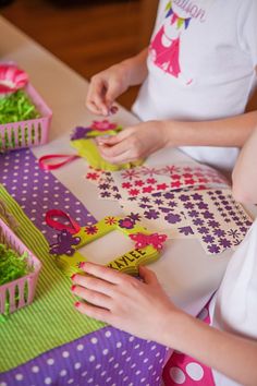 two girls are cutting paper with scissors on a purple and green table cloth, while another girl is wearing a white t - shirt