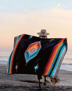 a woman is walking on the beach with a colorful blanket over her head and hat