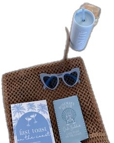 a beach towel with sunglasses and a book on it next to a drink cup in a straw bag