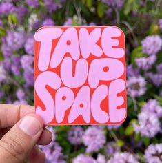 someone holding up a sticker that says take up space in front of purple flowers