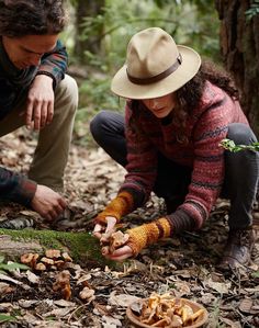 two people crouching down to look at something on the ground in the woods while one woman holds out her hand