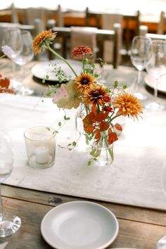 an arrangement of flowers in a glass vase on a table with plates and wine glasses