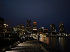 the city skyline is lit up at night with lights reflecting in the water and people walking on the sidewalk