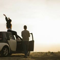 a man and woman standing next to a small car in the desert with their arms up