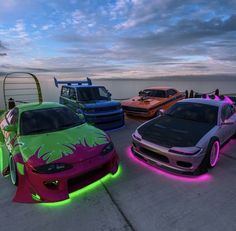 four different colored cars parked next to each other in front of the ocean at dusk