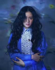 a woman with long black hair wearing a blue shirt and gold bracelets standing in front of a floral background