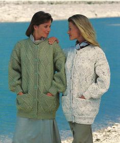 two women standing next to each other on the beach wearing green and white sweaters