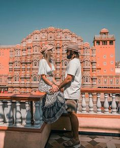 a man and woman sitting on a bench in front of a pink building with white balconies