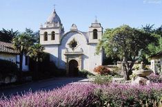 an old church surrounded by purple flowers and greenery in front of the entrance to it