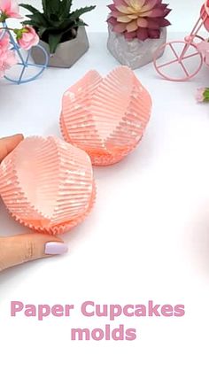 a hand holding two pink cupcake molds on top of a white table next to potted plants