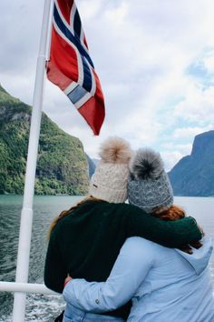 two people on a boat with a flag flying in the air over water and mountains