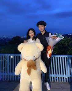 a man and woman standing next to a giant teddy bear with flowers in their hands