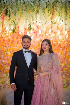 a man and woman standing next to each other in front of a floral wall with lights