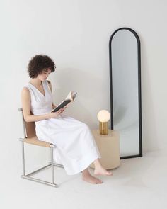 a woman sitting on a chair reading a book in front of a mirror and lamp