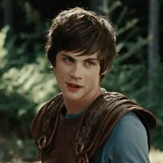 a young boy wearing a brown vest and blue shirt in front of some trees looking at the camera