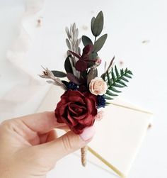 a hand holding a fake flower on top of a piece of paper with greenery