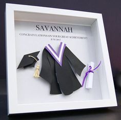 an origami graduation cap, gown and tassel in a white shadow box