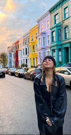 a woman standing in the middle of a street with cars parked on both sides and multi - colored buildings behind her