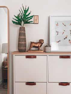 a white dresser topped with drawers and a vase filled with green plants next to a mirror