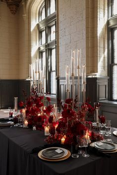 the table is set with candles, plates and red flowers in front of large windows