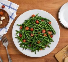 a white plate topped with green beans and tomatoes next to silverware on a wooden table