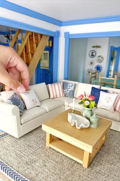 a hand is pointing at a dollhouse living room with blue walls and white furniture