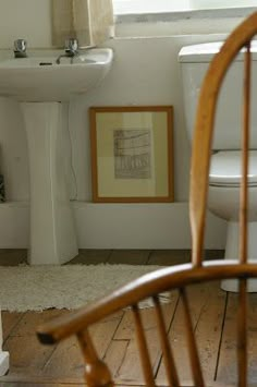 a white toilet sitting next to a sink in a bathroom under a window with a wooden chair