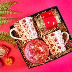 a red and white box with two mugs in it next to some flowers on a pink surface