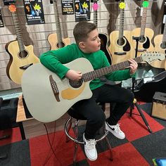 a young boy sitting on a stool playing an acoustic guitar in a music store with guitars behind him