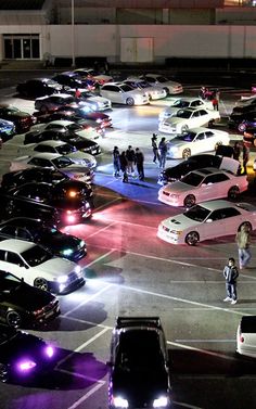 a parking lot filled with lots of parked cars and people walking around it at night