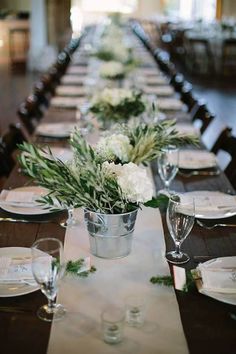 a long table is set with white flowers and greenery in buckets for centerpieces