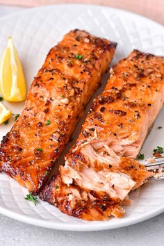 two pieces of salmon on a white plate with a fork and lemon wedges next to it