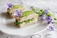 two sandwiches with cucumbers and herbs on a white plate next to purple flowers