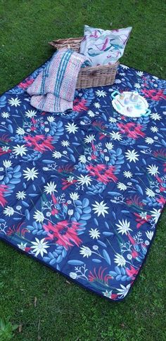 a blue blanket with flowers on it sitting in the grass next to a basket and pillows