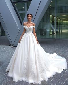 a woman standing in front of a building wearing a wedding dress with an off the shoulder neckline