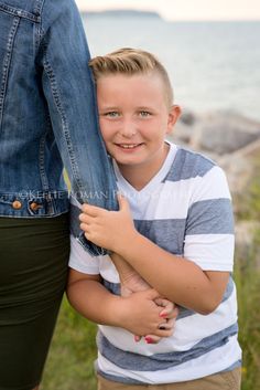 a young boy is hugging his mom's leg on the grass by the ocean