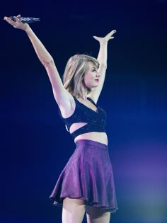 a woman in a purple skirt holding a microphone up to the air with her right hand