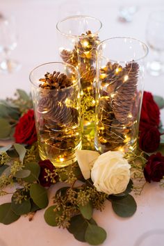 three glasses filled with pine cones on top of a table covered in flowers and greenery