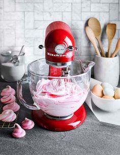 a red kitchen aid mixer with pink icing in the bowl next to eggs and utensils