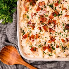 a casserole dish with bacon, cheese and parsley on the side next to a wooden spoon