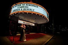 a couple standing in front of a marquee at night with their arms around each other