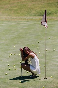 a woman kneeling down to pick up some tennis balls on the green with a flag in the background