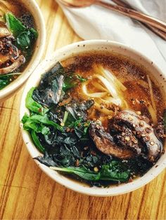 two bowls filled with soup and greens on top of a wooden table next to utensils