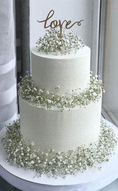 a three tiered cake with white frosting and flowers on the top is decorated with gold lettering that says love