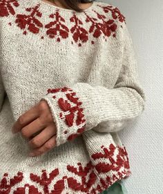 a woman wearing a sweater with red and white flowers on the sleeves, standing in front of a wall