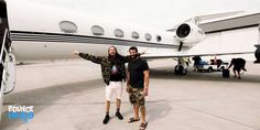 two men standing in front of an airplane with their arms out and one man pointing at the camera