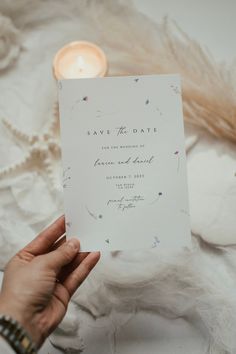 a person holding up a wedding card in front of a white background with flowers and feathers