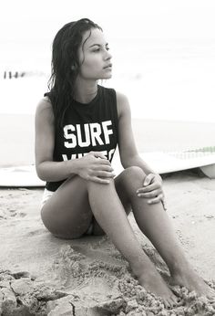 a woman sitting on the beach with her legs crossed and wearing a shirt that says surf