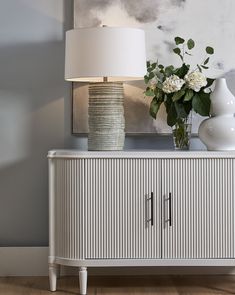 a vase with flowers sitting on top of a white cabinet next to a lamp and painting